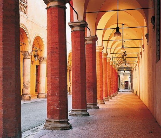 Un lungo portico con una fila di colonne ottagonali in mattoni rossi, un soffitto a volta e un'illuminazione calda da lampade a sospensione.