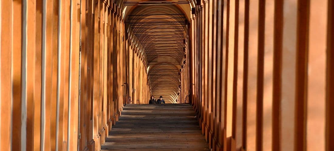 Due persone sono sedute su dei gradini in lontananza, lungo un lungo portico ad arco con colonne di terracotta e ombre che si proiettano dalla luce del sole.