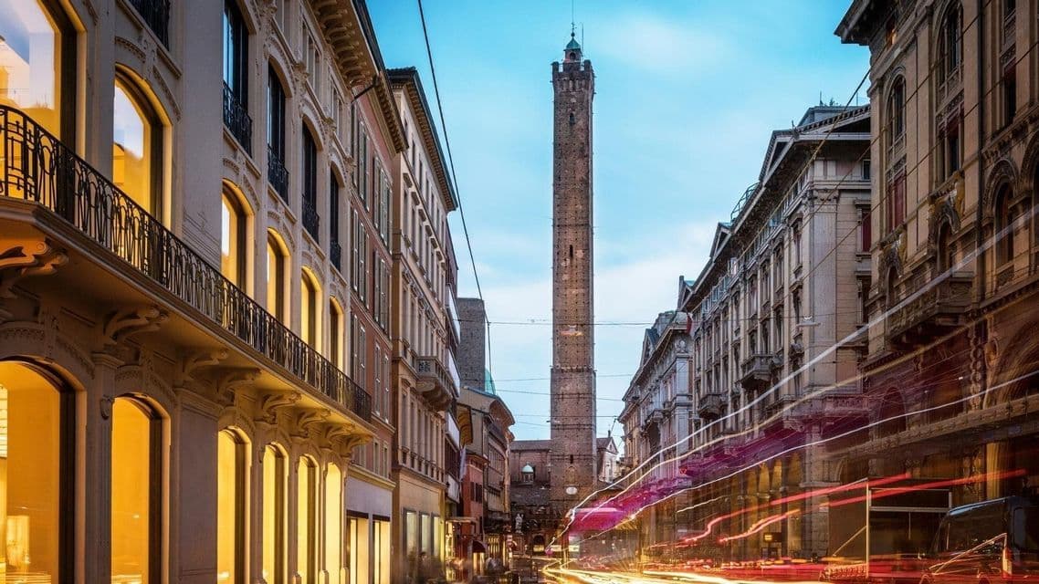 Una torre di mattoni alta si erge alla fine di una strada cittadina con edifici storici e scie luminose del traffico al crepuscolo.