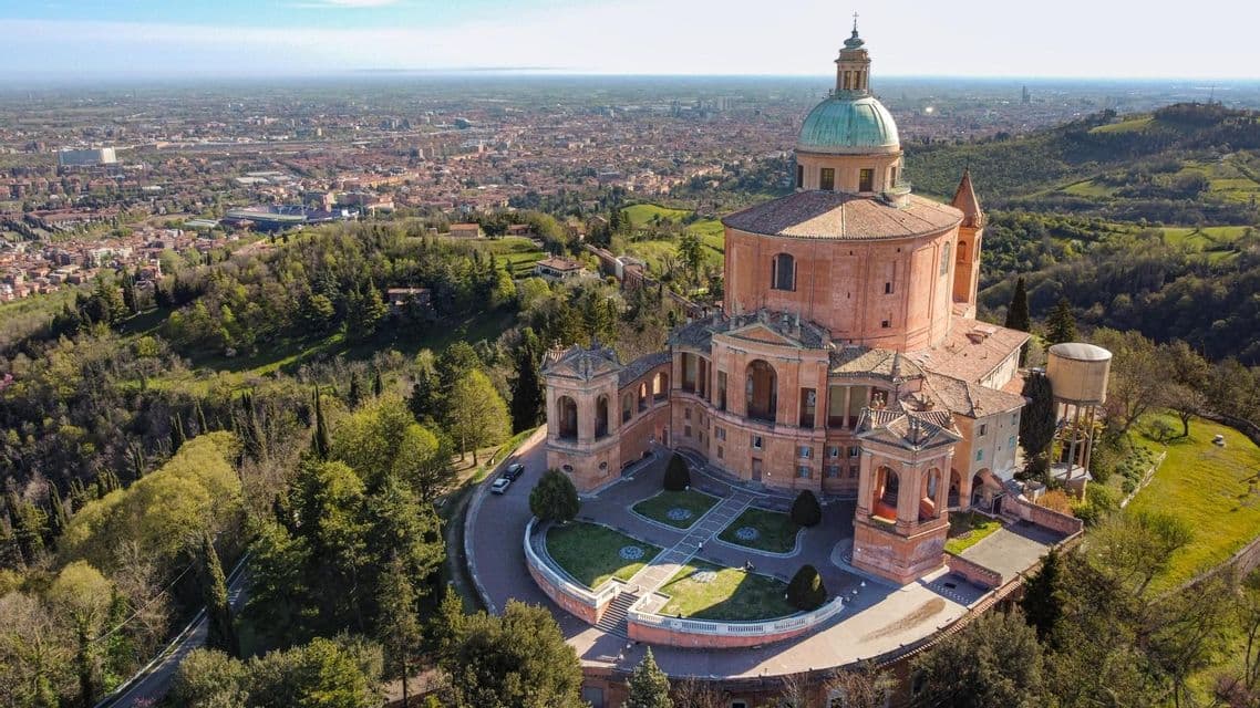 Veduta aerea di un santuario storico con una grande cupola, arroccato su una collina verde che domina un'ampia città.