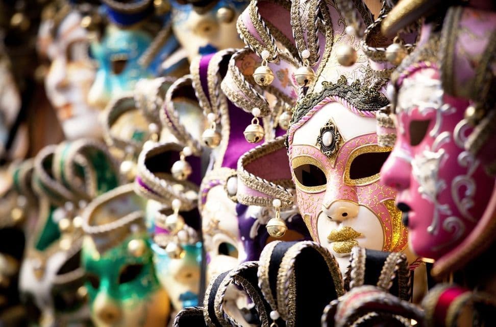 Des masques de carnaval ornés et colorés, décorés de liserés dorés et de clochettes, sont suspendus en rang, avec un masque rose et blanc au premier plan.