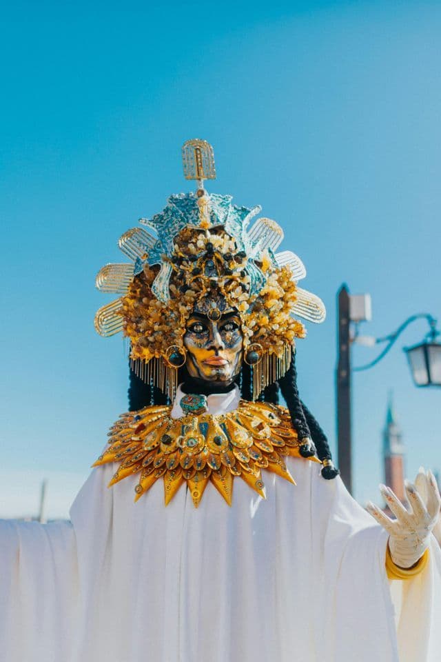 Une personne en costume élaboré avec un visage doré, une coiffe complexe bleue et dorée, et une robe blanche sous un ciel bleu clair.