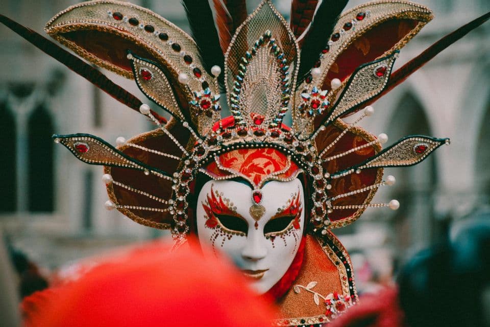 Un gros plan sur un masque de carnaval orné, rouge, blanc et or, avec une grande coiffe ornée de bijoux et de plumes.