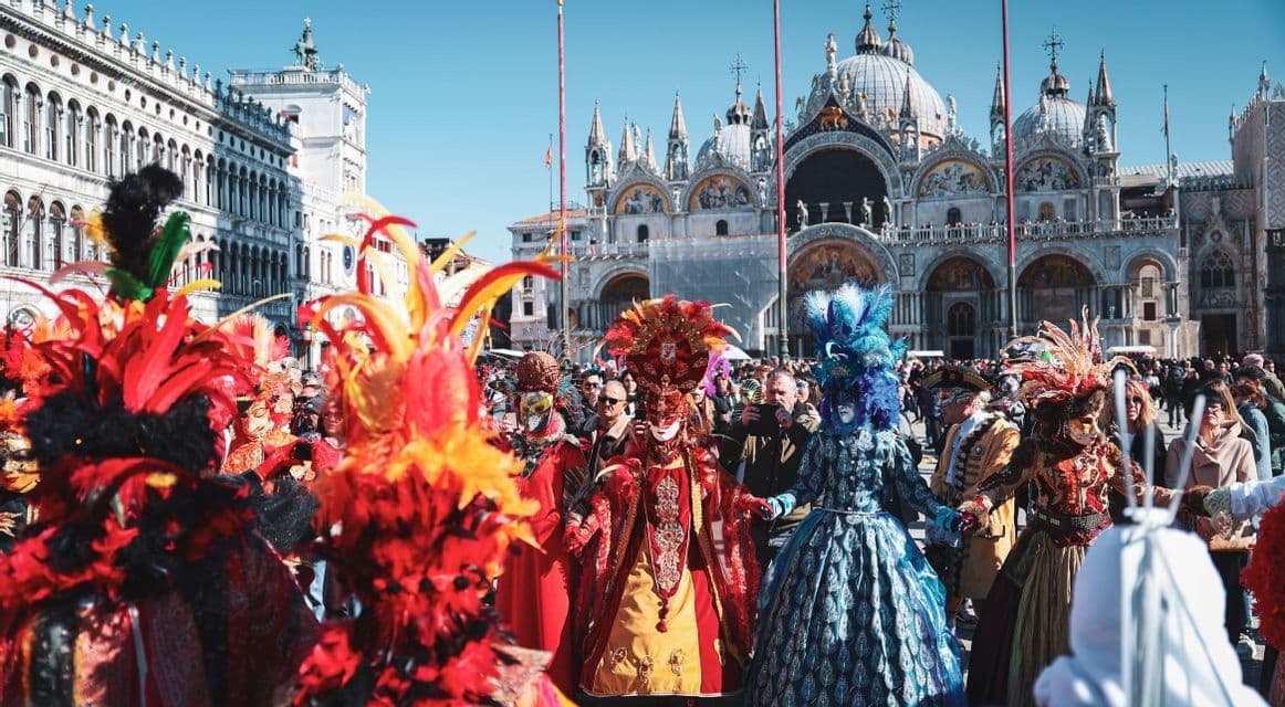 Une foule de personnes portant des masques de carnaval élaborés et colorés et des costumes à plumes célèbrent sur une place historique.