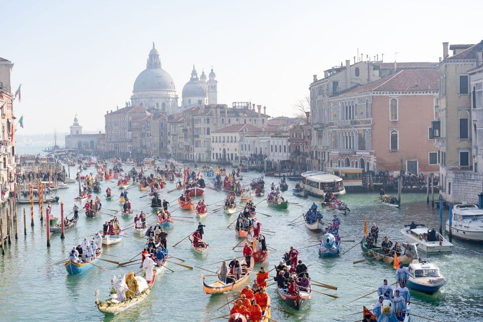 Des dizaines de barques colorées remplies de personnes costumées défilent sur un large canal, avec des bâtiments historiques et un dôme en arrière-plan.