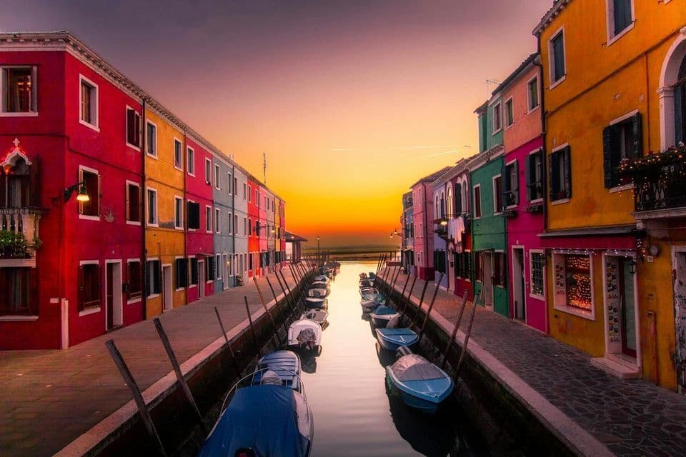 Rows of colorful buildings flank a narrow canal filled with moored boats under a glowing sunset sky.
