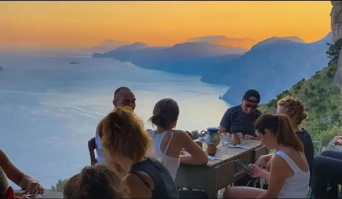 Un viaje en grupo de WeRoad se reúne alrededor de una mesa en un acantilado, con vistas a un vasto mar y una cadena montañosa al atardecer.