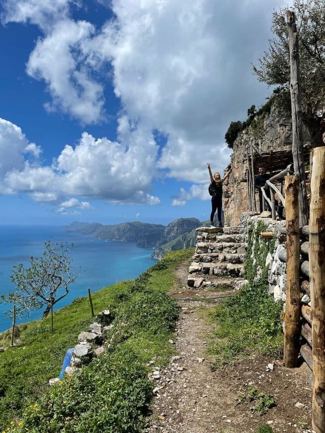 Una mujer de un viaje en grupo de WeRoad posa en un sendero junto al acantilado, con vistas a un mar turquesa y una costa montañosa.