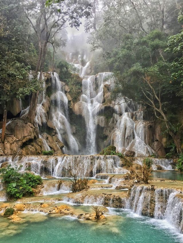 Una cascata a più livelli precipita lungo sporgenze rocciose in pozze turchesi, circondata da una foresta lussureggiante e nebbiosa.