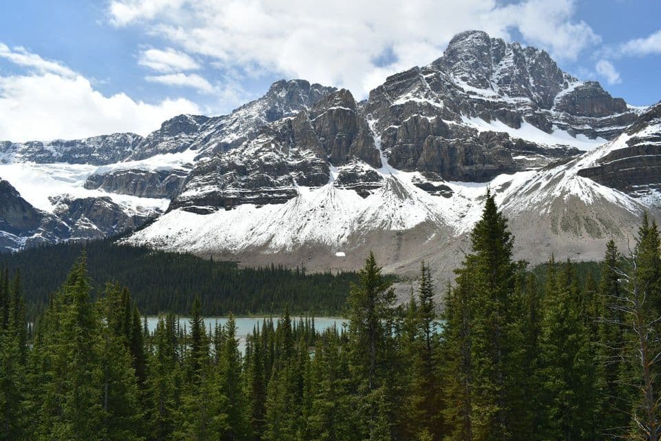 A dense pine forest stands before a turquoise lake at the base of large, snow-capped mountains under a cloudy sky.