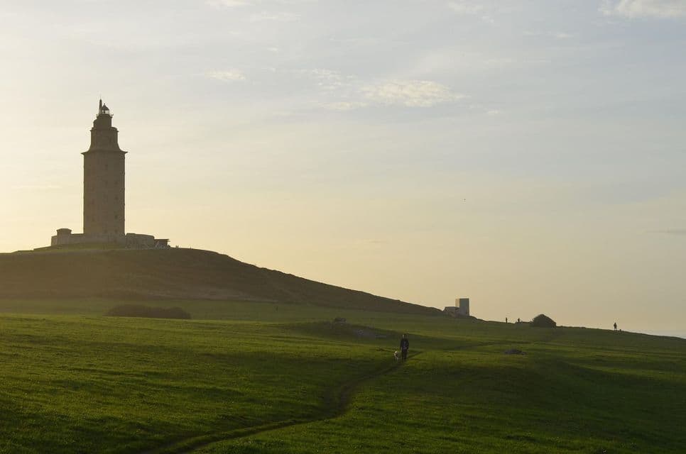 Un faro de piedra se recorta en una colina cubierta de hierba contra un cielo pálido al atardecer, con una persona y un perro caminando por un sendero debajo.