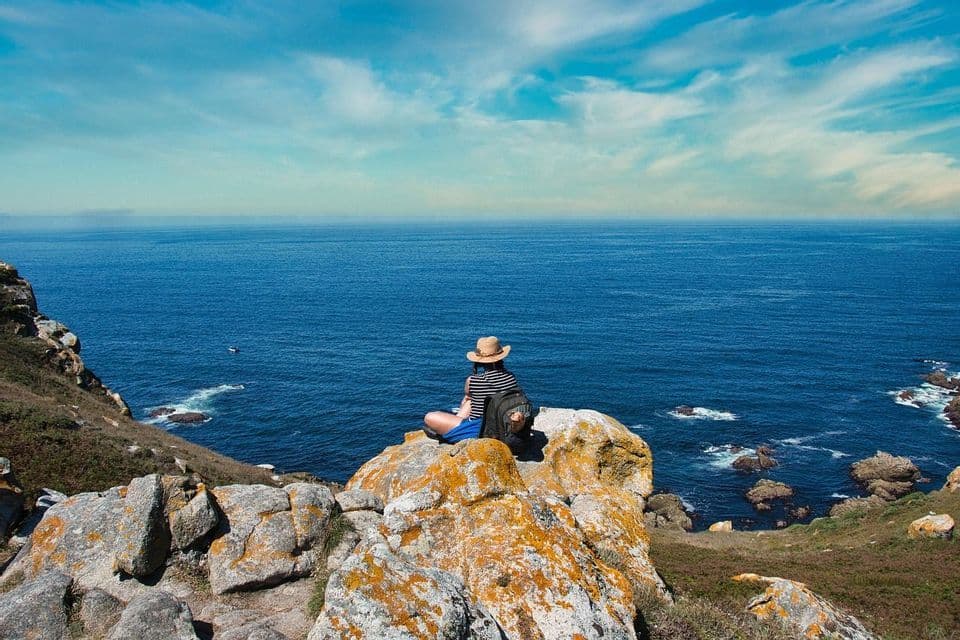 Una persona con un sombrero de paja y una mochila se sienta en un acantilado rocoso con vistas al vasto océano azul bajo un cielo parcialmente nublado.