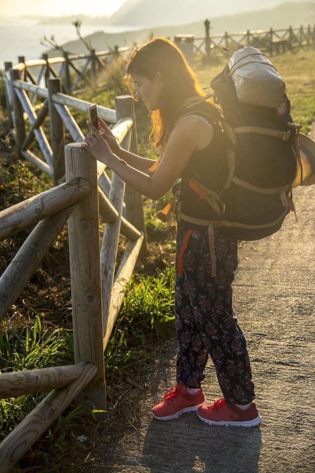 Una mujer con una gran mochila de senderismo se apoya en una valla de madera y toma una foto con su smartphone al atardecer.