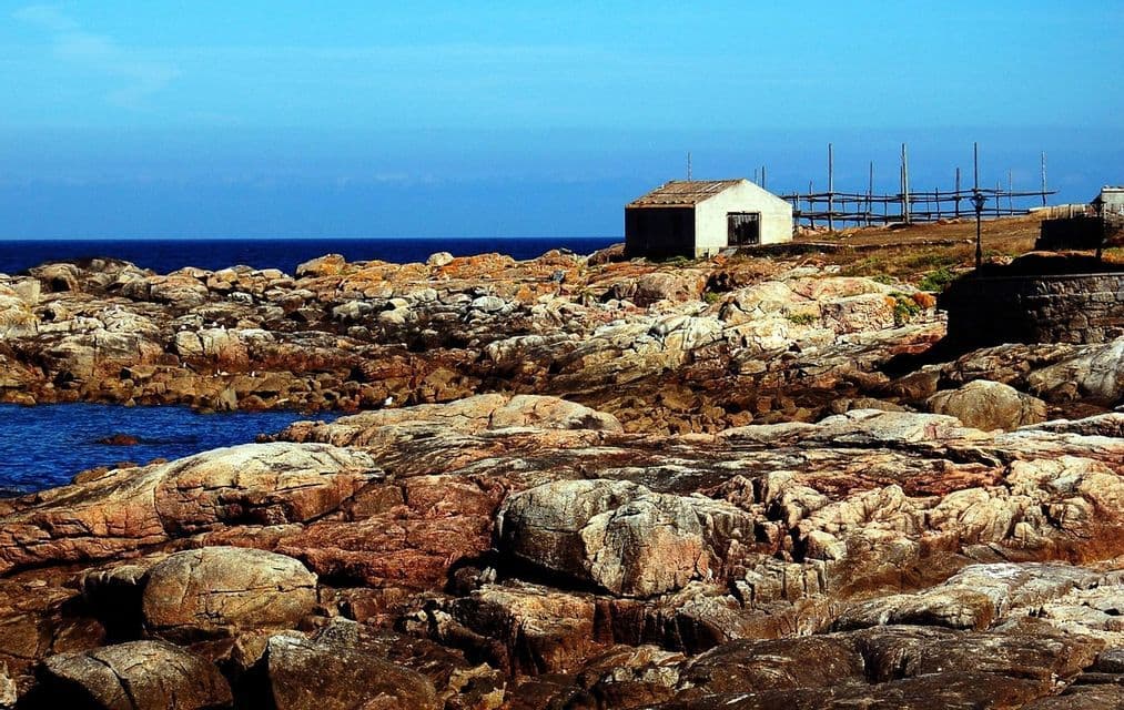 Un pequeño edificio blanco se encuentra en una costa escarpada y rocosa, junto al mar azul profundo bajo un cielo despejado.