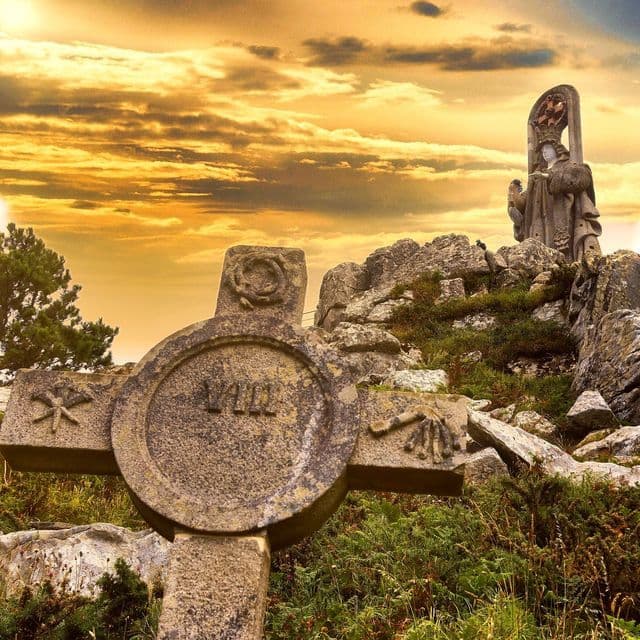 Una cruz de piedra tallada y una estatua se alzan en una ladera rocosa y verde contra un dramático cielo dorado al atardecer.