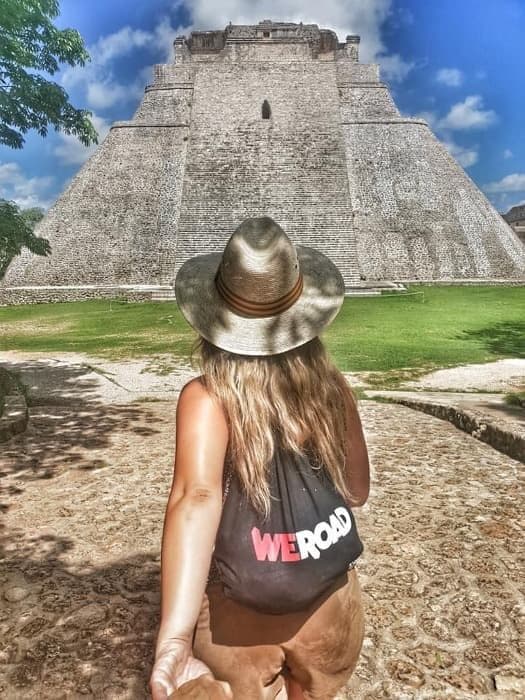 A woman on a WeRoad group trip, seen from behind, walks towards a large stone pyramid on a sunny day.