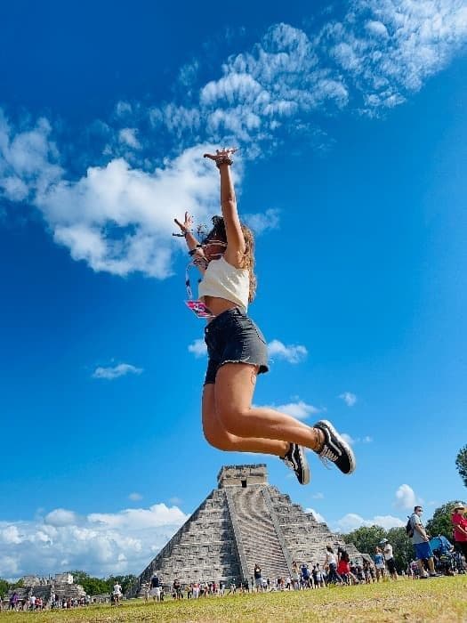 A woman jumps high in the air in front of a large stone pyramid, with a crowd of people below on the grass under a blue sky.
