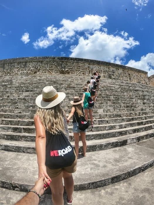 A person holds a woman's hand as she and a WeRoad group trip climb the stone steps of a large ancient ruin under a blue sky.