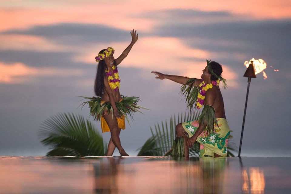 Zwei Personen in traditioneller polynesischer Kleidung mit Blumenketten tanzen bei Sonnenuntergang an einem Pool neben einer beleuchteten Tiki-Fackel.