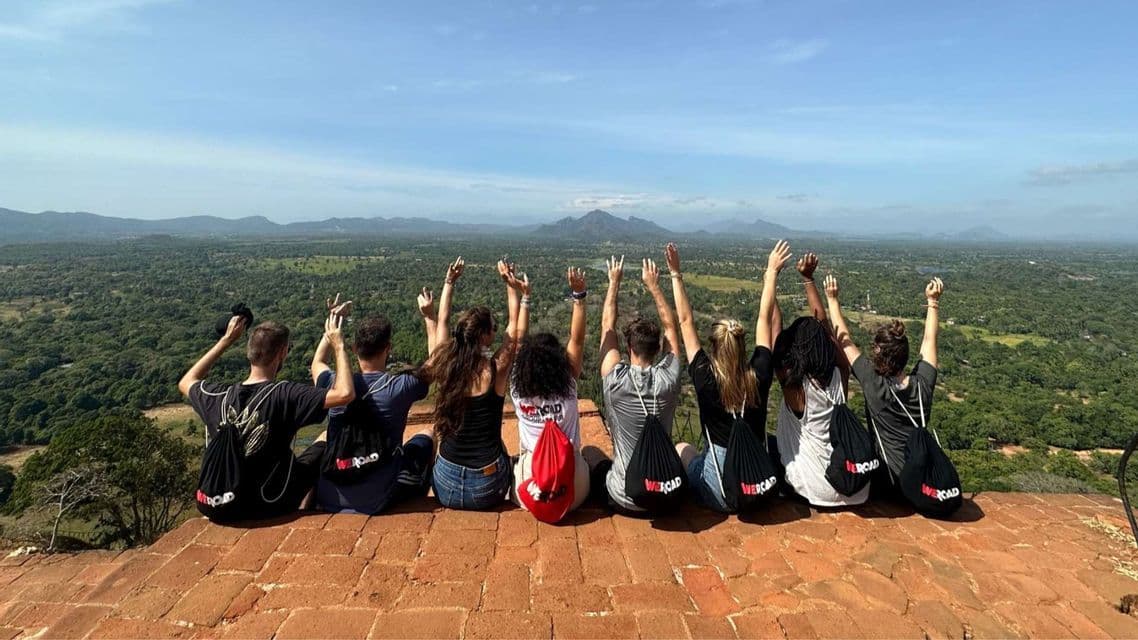 Un gruppo WeRoad, con le spalle alla fotocamera e le braccia alzate, ammira una vasta valle verde e le montagne da un punto panoramico elevato.