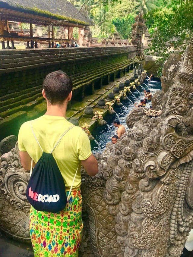 Eine Person mit einem WeRoad-Rucksack und einem Sarong blickt auf badende Menschen unter geschnitzten Wasserspeiern in einem steinernen Tempelbecken.