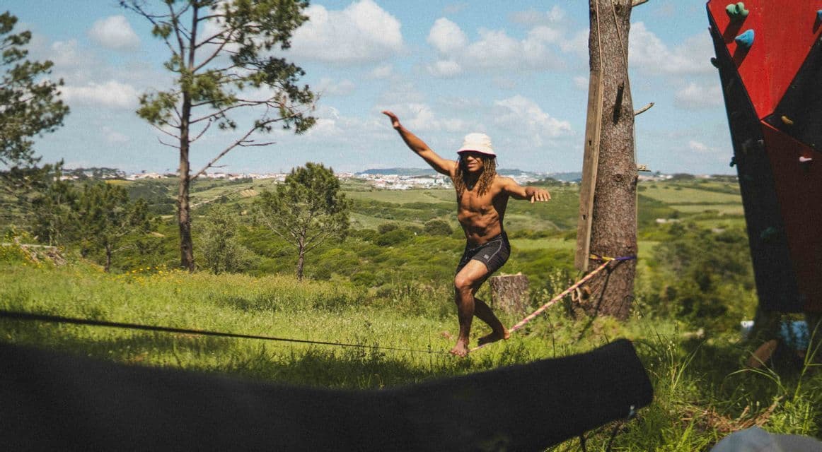 Una persona a torso nudo con un cappello da pescatore si bilancia a piedi nudi su una slackline in un paesaggio erboso e collinare sotto un cielo parzialmente nuvoloso.