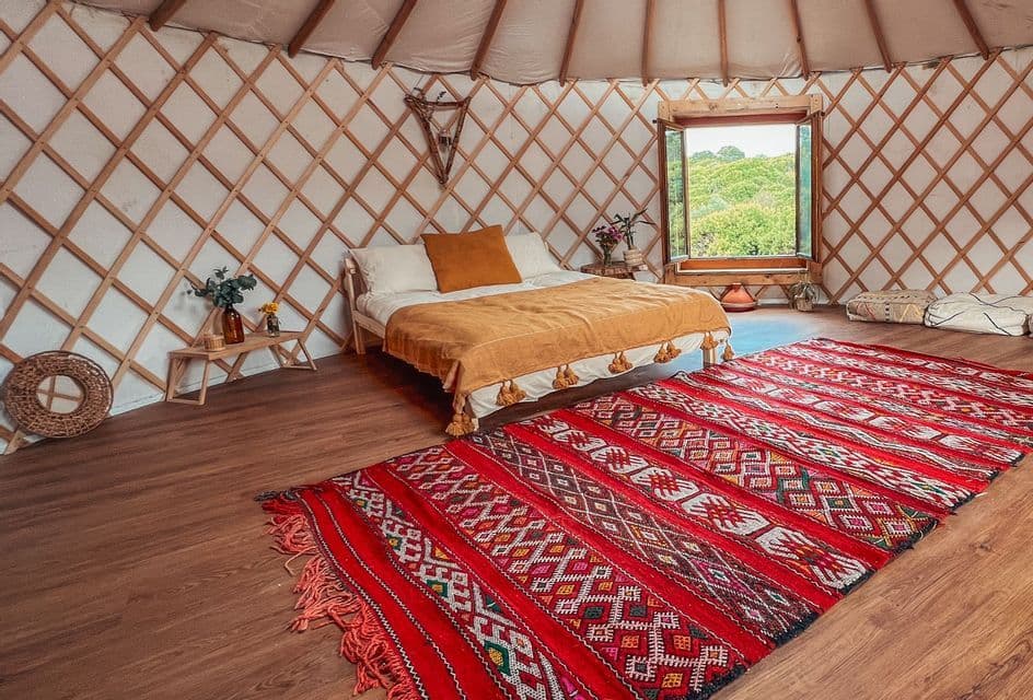 L'interno di una yurta con un letto fatto, un grande tappeto rosso a motivi su un pavimento di legno e una finestra che si affaccia su colline verdi.