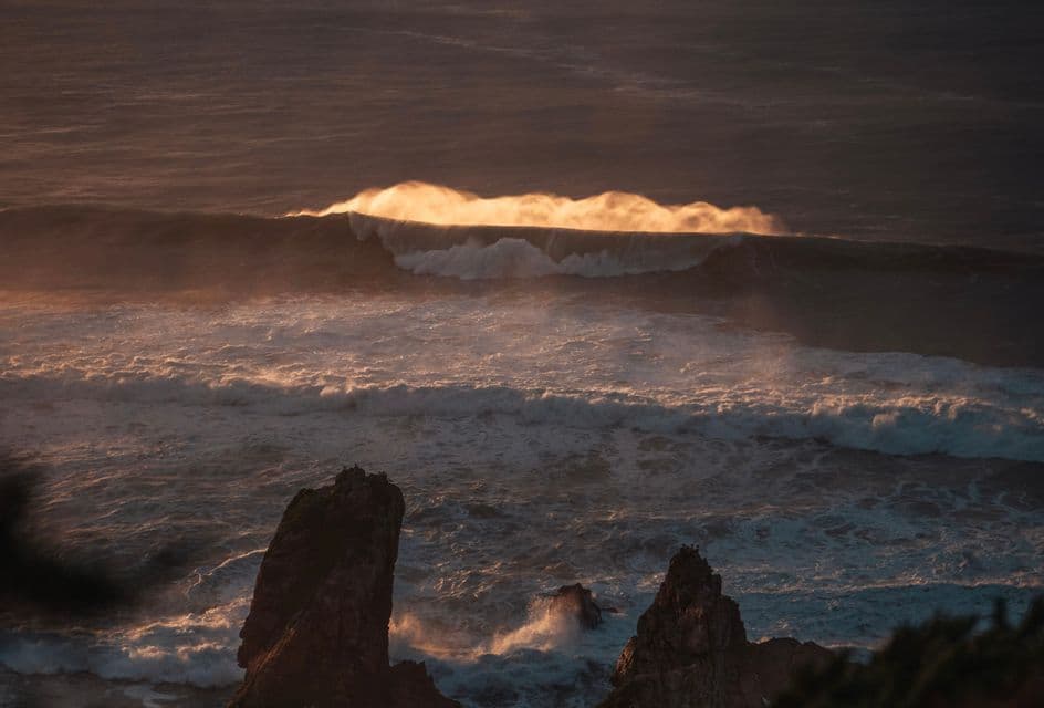 Una grande onda oceanica illuminata dalla luce dorata del tramonto si dirige verso formazioni rocciose nel mare.