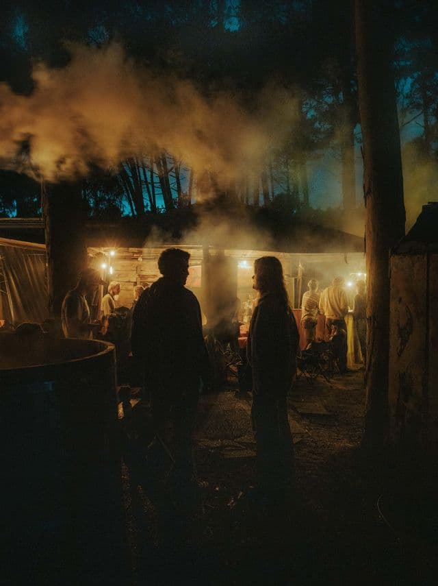 Un gruppo WeRoad si ritrova in un rifugio calorosamente illuminato in una foresta di notte, con due persone in silhouette in primo piano.