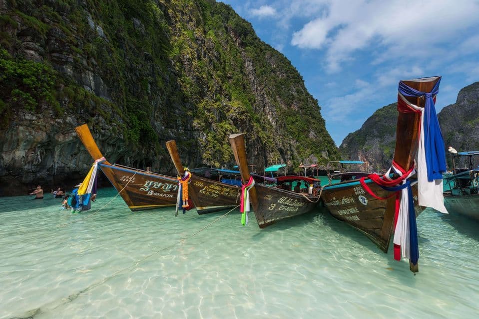 Plusieurs bateaux traditionnels à longue queue amarrés dans des eaux peu profondes, claires et turquoise, à côté d'une falaise rocheuse luxuriante.