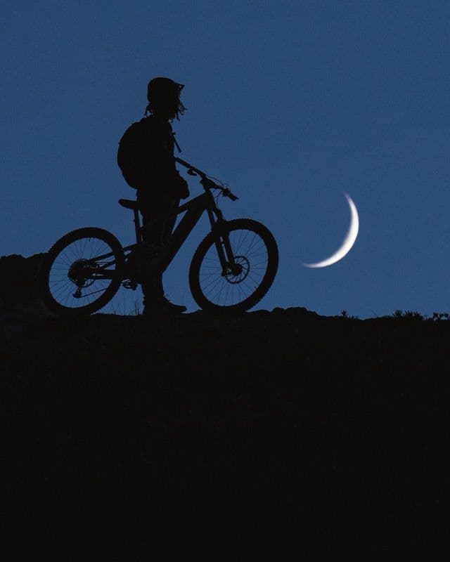 Un ciclista in silhouette con una mountain bike si erge su una collina contro un cielo blu scuro con una luna crescente.