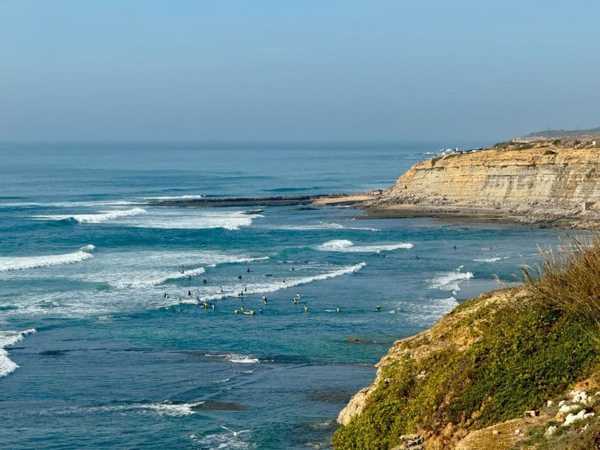 Una veduta dall'alto di numerosi surfisti nell'oceano blu con onde bianche, accanto a una scogliera rocciosa.