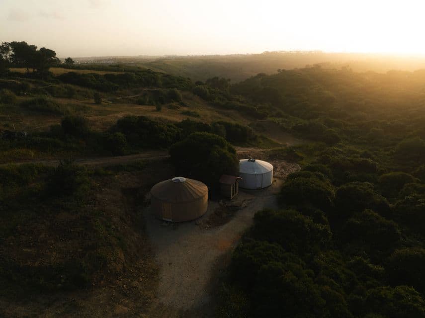 Vista aerea di due yurte su una radura sterrata immersa in dolci colline verdi sotto un cielo sfumato al tramonto.