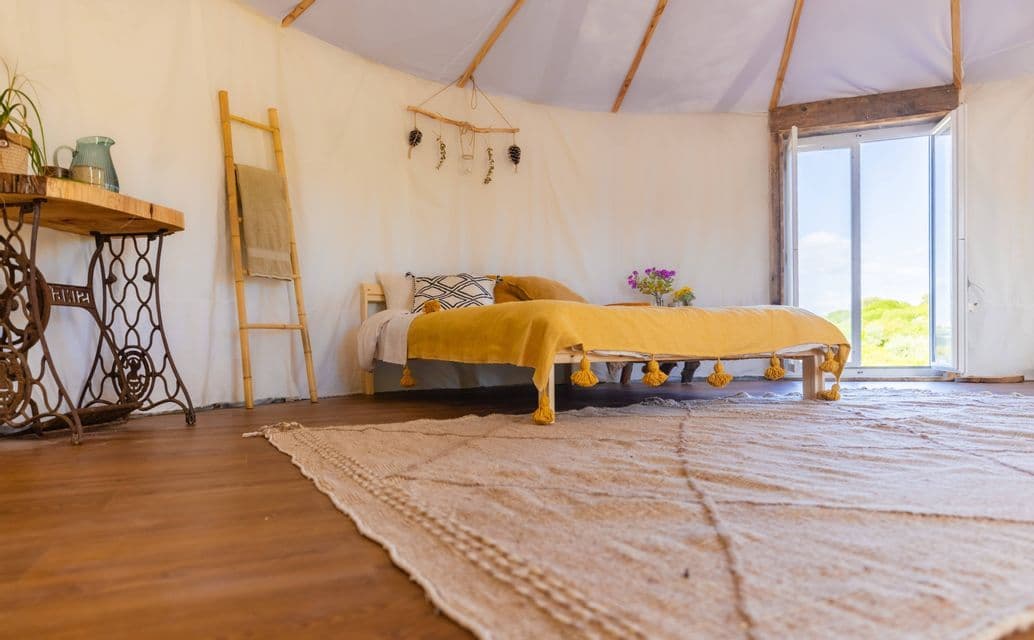 Interno rustico di una tenda stile yurta con letto basso, coperta gialla, tappeto intrecciato e porta aperta.