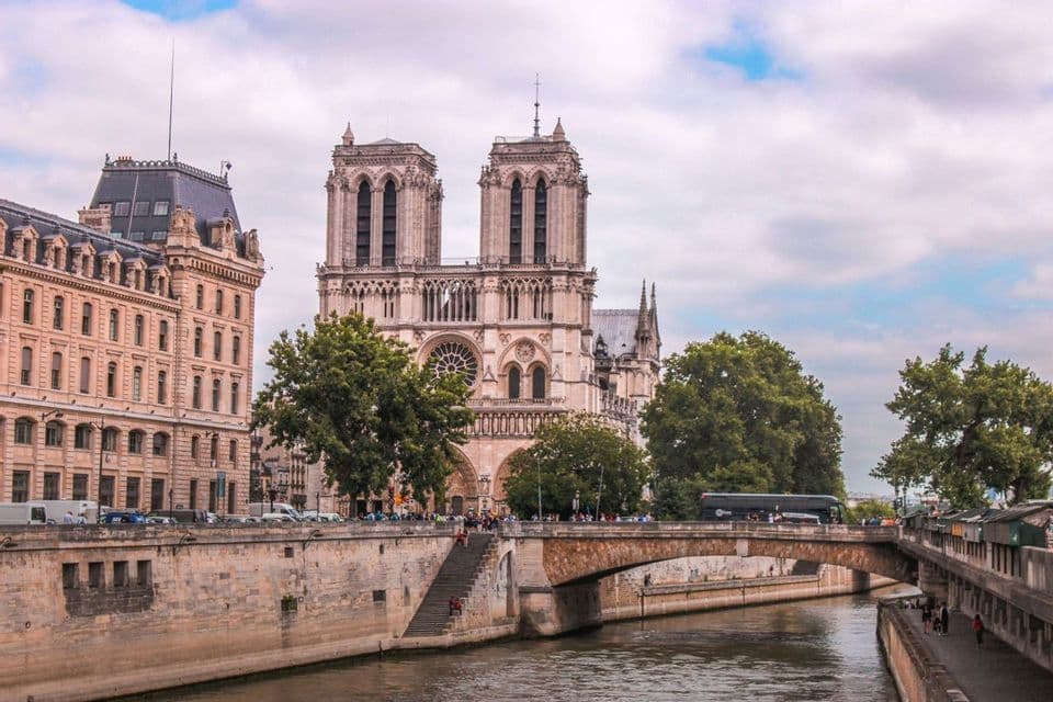 Una gran catedral histórica con dos torres se alza a orillas de un río, vista desde el otro lado del agua, pasando un puente de piedra.