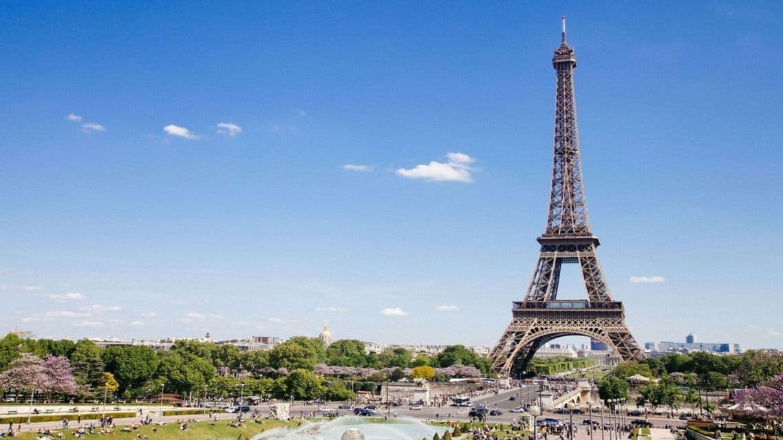 La Torre Eiffel se alza contra un cielo azul con nubes dispersas, dominando un parque lleno de árboles verdes y gente.