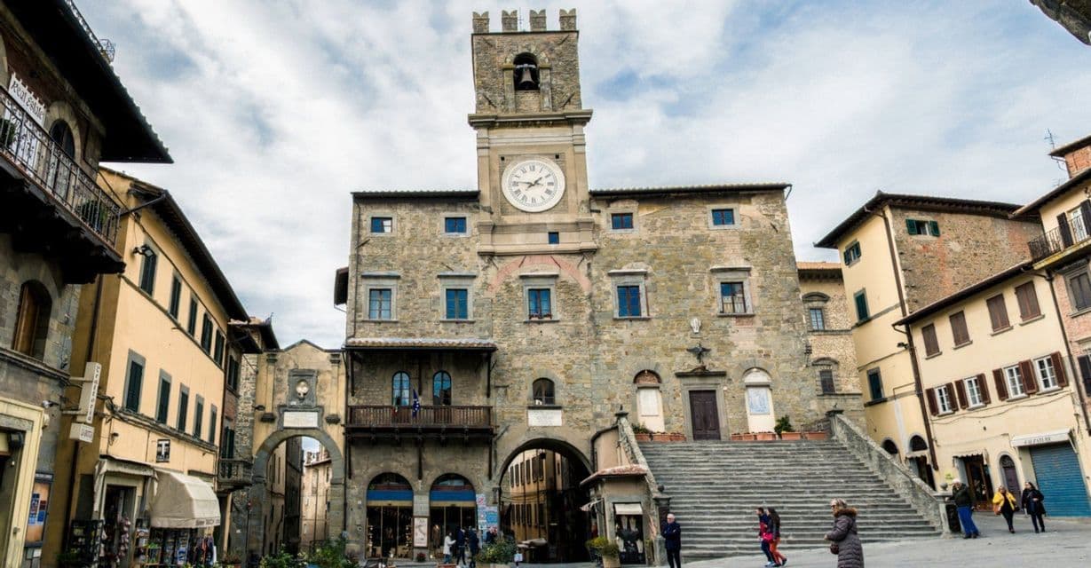 Un edificio in pietra con torre dell'orologio si erge in una piazza cittadina, con una grande scalinata e persone che passeggiano in una giornata nuvolosa.