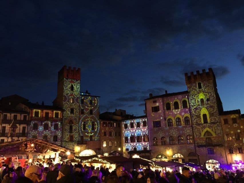 Motivi luminosi colorati proiettati su edifici storici sopra un affollato mercato notturno in una piazza cittadina.