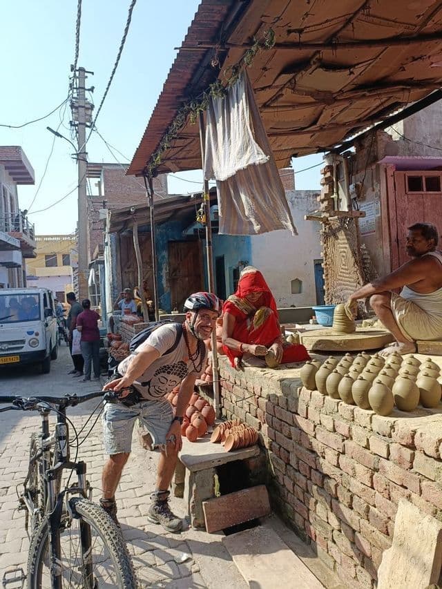 Un uomo in un viaggio di gruppo WeRoad con la sua bicicletta sorride mentre posa accanto a vasai locali che lavorano in una strada di paese.