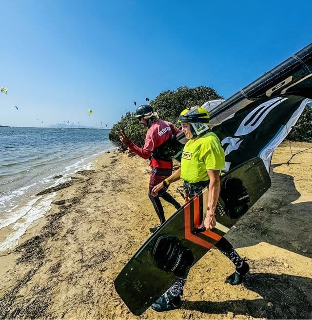 Dos personas de un viaje en grupo de WeRoad, con cascos y trajes de neopreno, caminan por una playa de arena llevando equipo de kitesurf.