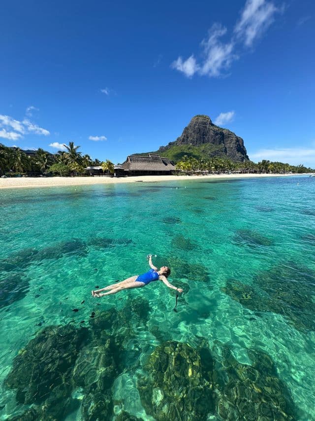 Una mujer en traje de baño azul flota de espaldas en agua cristalina turquesa, con una playa tropical y una gran montaña al fondo.