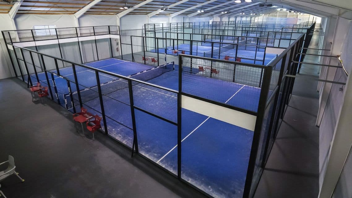 Vista dall'alto di campi da padel blu vuoti con pareti in vetro disposti in file all'interno di una grande struttura sportiva al coperto.