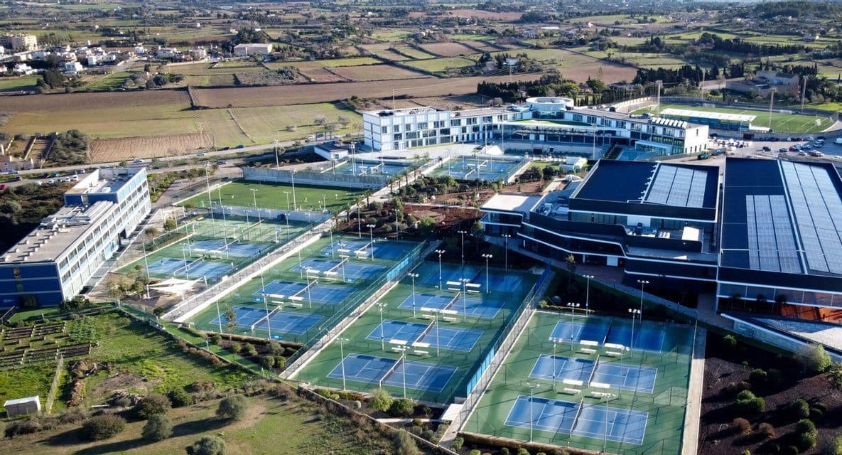 Una vista aerea di un moderno complesso di accademia di tennis con numerosi campi blu e verdi, circondato da edifici e un paesaggio rurale.