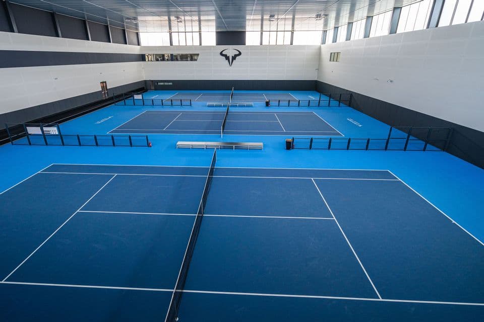 Diversi campi da tennis azzurri e vuoti con reti, all'interno di un grande impianto sportivo al coperto.