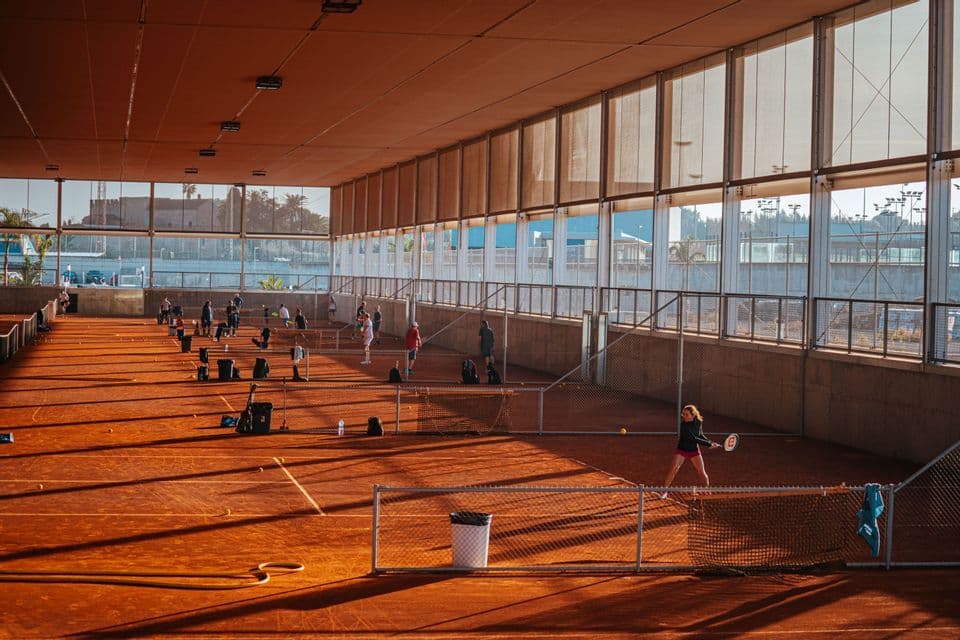 Diverse persone giocano a tennis su campi indoor in terra rossa, con la luce del sole intensa che crea lunghe ombre.