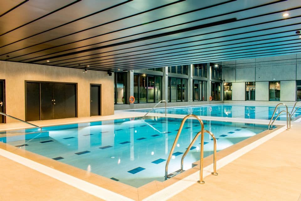 Una piscina interna moderna e vuota con acqua blu luminescente, scalette in acciaio inossidabile e un soffitto a doghe scure.