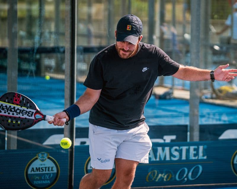 Un uomo con maglietta e cappellino neri che colpisce una palla con una racchetta da padel su un campo blu.