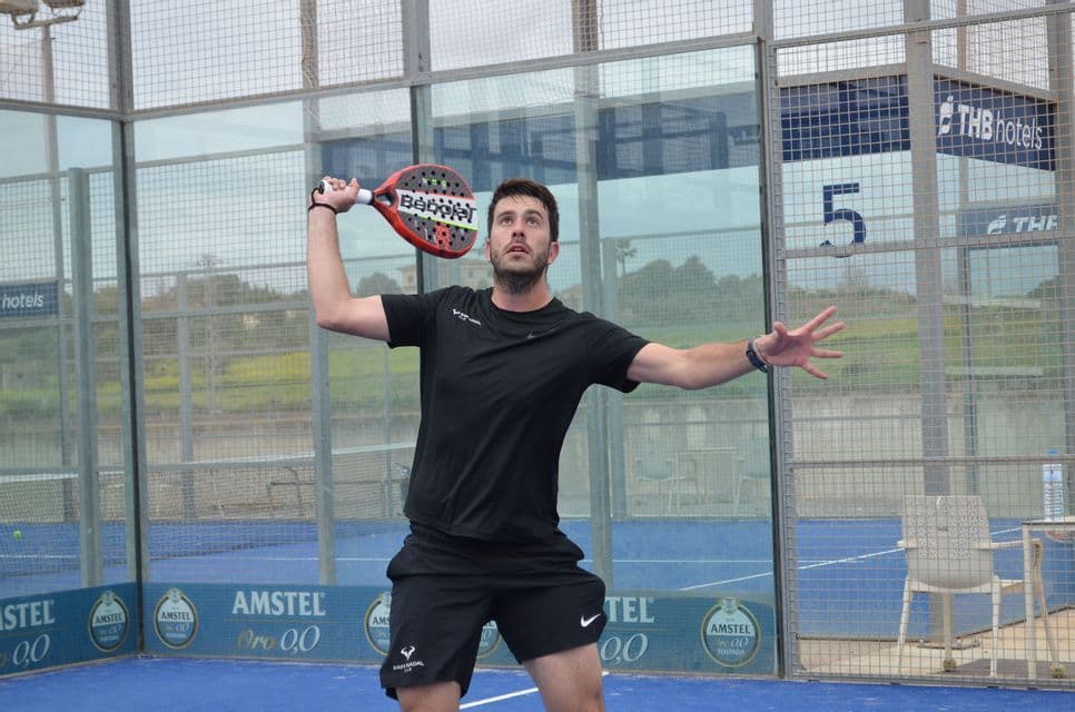 Un uomo in abbigliamento sportivo nero alza la sua racchetta da padel rossa, preparandosi a servire su un campo da padel blu recintato.