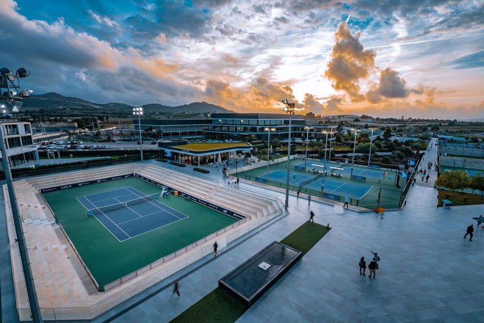 Un ampio complesso sportivo con numerosi campi da tennis ed edifici moderni, visto dall'alto durante un tramonto spettacolare.