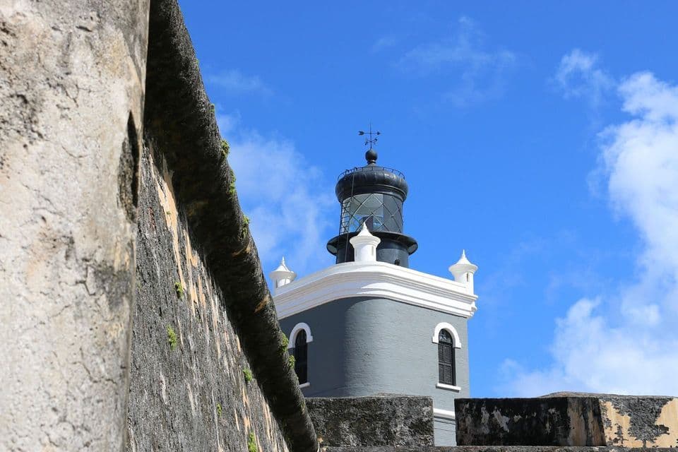 Veduta dal basso di un faro grigio con una lanterna nera, oltre un muretto in pietra eroso sotto un cielo azzurro.
